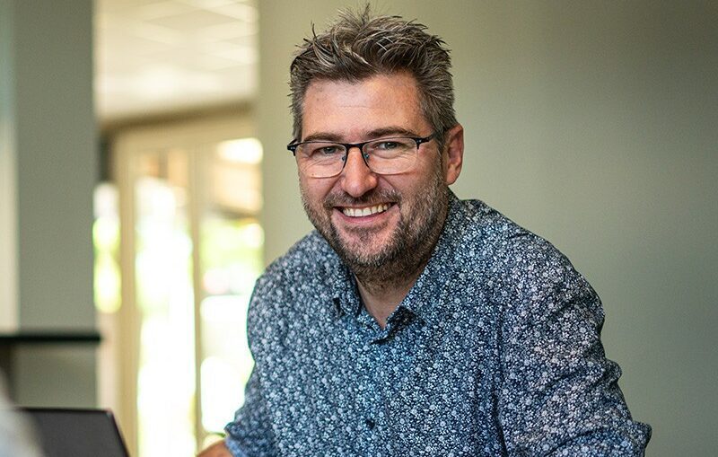 Homme souriant avec lunettes et chemise à motifs, assis dans un bureau lumineux avec un ordinateur portable.