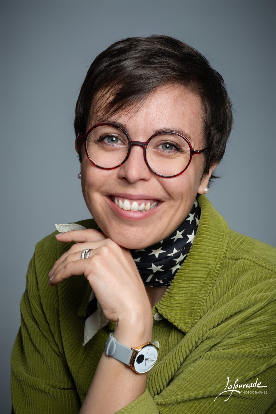 Femme souriante portant des lunettes rondes rouges, un pull vert et une echarpe noire a etoiles blanches