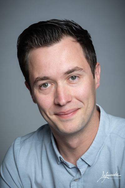 Portrait d'un homme souriant en chemise bleu clair, cadré en plan rapproché, avec un fond gris uni.