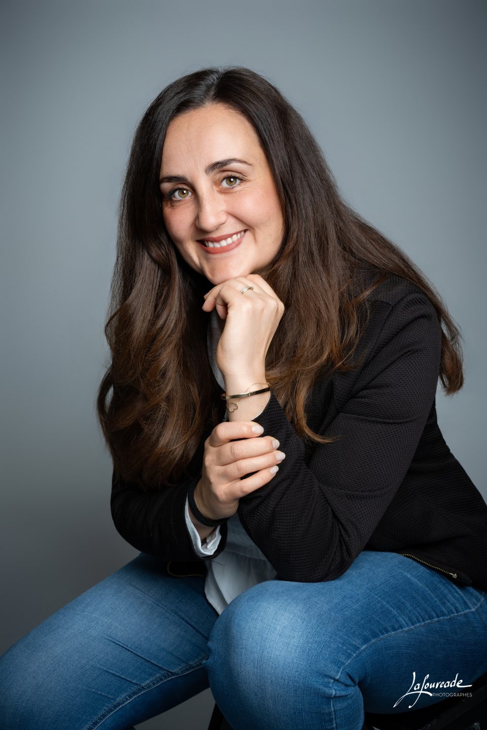 Femme souriante en veste noire et chemise blanche, posture décontractée, bijoux discrets, arriere-plan gris