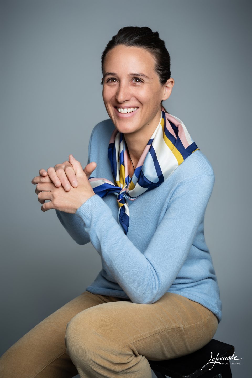 Femme souriante en pull bleu et foulard coloré, assise dans une posture décontractée.