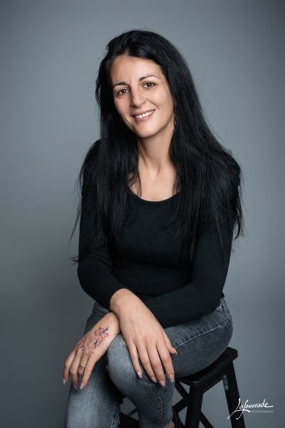 Femme souriante en haut noir et jean gris, assise sur un tabouret noir, avec un tatouage coloré sur la main.